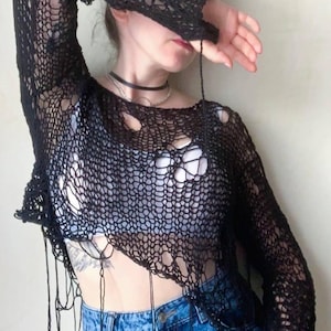May include: Black, open-weave long-sleeve top with a distressed design. The top features a loose, net-like pattern with intentional holes and dangling threads. Paired with blue denim jeans, the top creates a casual, edgy aesthetic.