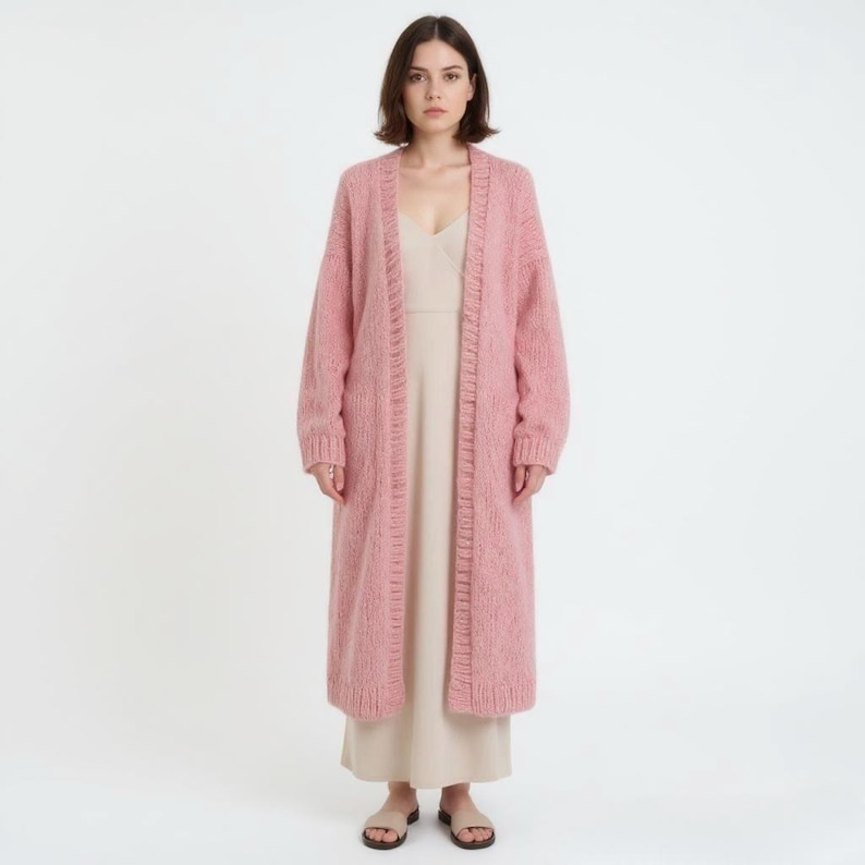 A woman poses in a fluffy, ankle-length pink cardigan and a beige dress.