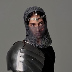 May include: A person wearing a gray chainmail coif with a silver headpiece and a silver shoulder piece of armor.