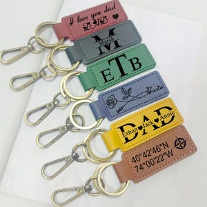 May include: A collection of personalized keychains in various colors, including pink, gray, green, blue, yellow, and brown. Each keychain features a rectangular leather tag with text such as "I love you dad," initials, names, and coordinates. The keychains have gold-colored rings and clasps.