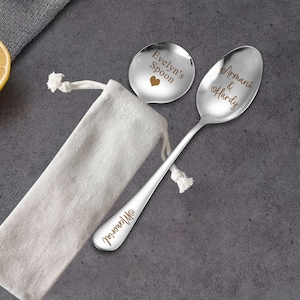 Può includere: Due cucchiai in metallo color argento con incisioni personalizzate. Un cucchiaio presenta il testo 'Evelyn's Spoon' con un'icona a forma di cuore, mentre l'altro mostra 'Armani & Hardy' con icone a cuore e la parola 'Memorial' incisa sul manico. Un sacchetto in tessuto beige.