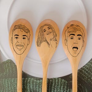 May include: Three wooden spoons with engraved portraits. Each spoon features a different person's face, rendered in black line art. The spoons are arranged on a white plate, suggesting a kitchen or dining setting. The spoons are made of light-coloured wood.