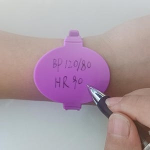 May include: A purple wristband with the text "BP 120/80 HR 90" written on it. A pen is held near the wristband, as if writing on it. The wristband is on a person's wrist.