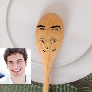 May include: A wooden spoon with a person's face engraved on the front. The face is smiling and has a beard. The spoon is light brown and has a natural wood grain.