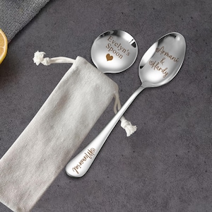 May include: Two silver spoons with personalized engravings. One spoon says "Evelyn's Spoon" with a heart and the other says "Armani & Hardy" with a heart. The spoons are on a white fabric surface.