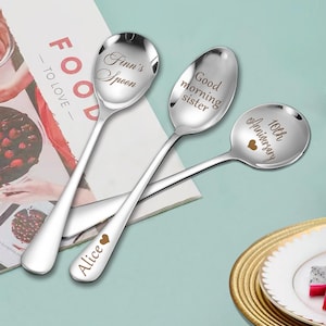 Op de afbeelding: Drie zilveren lepels met gegraveerde berichten. De lepels liggen op een wit oppervlak met een tijdschrift op de achtergrond. De berichten op de lepels zijn "Finn's Spoon", "Good morning sister" en "10th Anniversary".