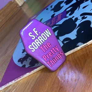 May include: A coffin-shaped pin with a metallic purple finish. The text S.F. SORROW is printed in white, and the text The Pretty Things is printed in pink. The pin is positioned over a vinyl record sleeve featuring a blue and black graphic design.