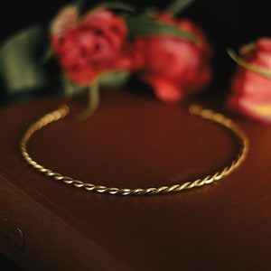 Könnte beinhalten: Eine goldfarbene Metall-Halskette im gedrehten Seildesign. Die Halskette liegt auf dem Einband eines dunkelbraunen Buches, platziert vor drei roten und rosafarbenen Tulpen mit grünen Stielen und Blättern vor einem schwarzen Hintergrund.