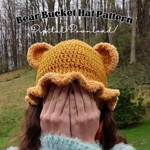 May include: A crochet bucket hat pattern with bear ears. The hat is yellow with a brown trim. The pattern is a digital download.