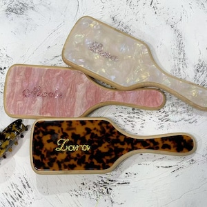 Op de afbeelding: Drie haarborstels met gepersonaliseerde namen in strass. De borstels hebben verschillende patronen: roze met witte swirls, wit met iriserende swirls en bruin met een schildpadpatroon.