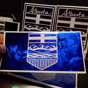Può includere: Una collezione di adesivi a tema Alberta. L'adesivo principale presenta uno sfondo blu con un emblema bianco di montagne, grano e una croce. La parola "Alberta" è scritta in corsivo. Un righello con "StickUp.ca" è in basso.
