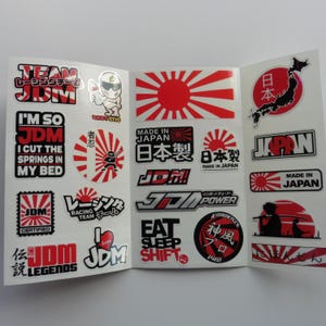 May include: A sticker sheet featuring various JDM-themed designs in red, black, and white. Graphics include the Japanese rising sun flag, text such as 'JDM Legends', 'Eat Sleep Shift', 'Made in Japan', and 'I'm so JDM', along with a map of Japan and a samurai silhouette.