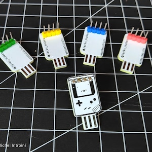 Puede incluir: Una colección de memorias USB de colores y una memoria USB con forma de consola de juegos retro. Las memorias USB tienen cuerpos blancos con tapas de colores y conectores USB. La memoria de la consola de juegos es en blanco y negro.