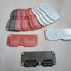 May include: A stack of red and white circuit boards with copper tracks and holes for soldering components. The boards are arranged in a fan shape.