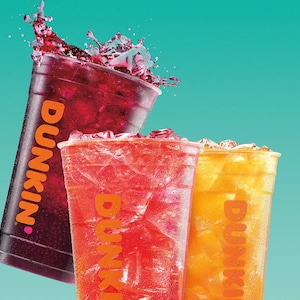 May include: Three iced beverages in clear plastic cups with the word "Dunkin'" printed on them. One is a dark red color with a splash, another is red, and the third is yellow. The background is a teal color.