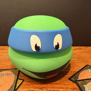 May include: A 3D-printed green Teenage Mutant Ninja Turtle head with a blue headband, white eyes, and a black smile. The figure is sitting on a wooden surface with Magic: The Gathering cards.