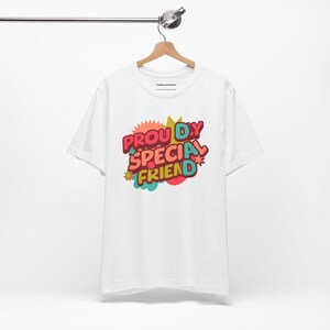 Pode incluir: Camiseta branca com um gráfico colorido que diz "Proudly Special Friend" em uma fonte retrô.