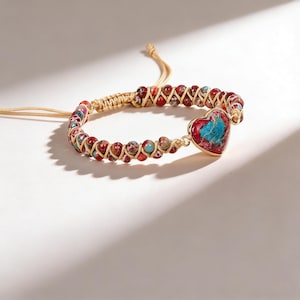May include: A handmade bracelet featuring a heart-shaped pendant with red and turquoise stone inlay. The bracelet is made with red, brown, and tan beads, and a woven cord. The heart pendant is framed in gold-colored metal.