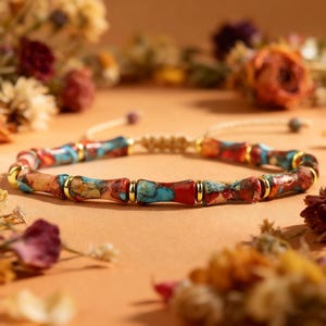 May include: A beaded bracelet with a macrame closure. The bracelet features cylindrical beads in shades of red, blue, and brown, separated by gold-colored metal accents. The background is a warm, neutral tone with floral accents.