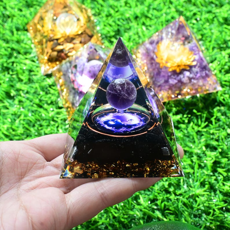 May include: A transparent pyramid with a purple sphere inside, set against a green background. The pyramid has a black base with gold flakes and a purple sphere suspended in the centre. Other pyramids with gold and purple accents are visible.