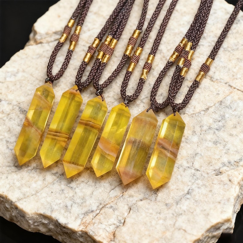 May include: Six necklaces with yellow and orange crystal pendants. The pendants are rectangular with pointed ends and are suspended from brown braided cords. The cords have gold-colored metal accents near the top. The necklaces are displayed on a light-colored stone surface.