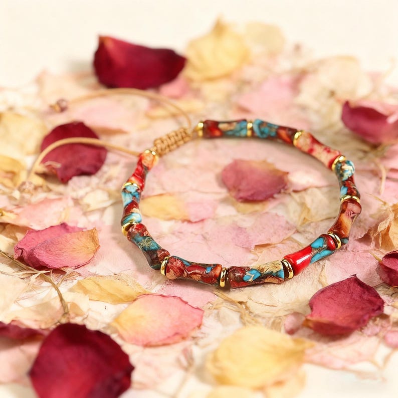 May include: A colorful beaded bracelet with red, blue, and gold accents. The bracelet is made of cylindrical beads and is set against a backdrop of rose petals. The bracelet is adjustable with a woven cord.