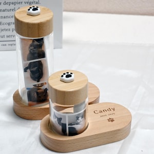 Op de afbeelding: Twee houten potten met glazen inzetstukken, elk met een reeks foto's van katten. De potten hebben deksels met pootjes en staan op houten bases. De basis van de pot rechts heeft de tekst "Candy 2012-2025" erop gegraveerd.