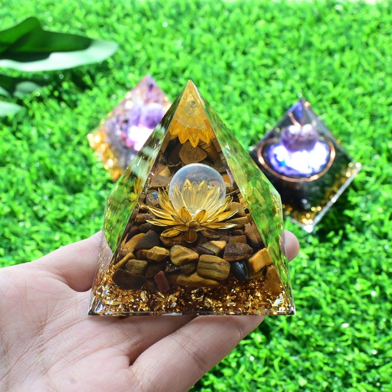 May include: A clear pyramid-shaped resin art piece with a golden lotus flower and a clear sphere inside. The pyramid is filled with brown stones and gold flakes. Two other pyramids are in the background, one purple and one with a blue centre.
