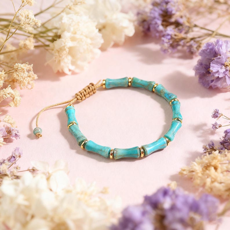 May include: Turquoise bamboo-style bracelet with gold-colored accents. The adjustable bracelet features light blue beads and a woven beige cord. The jewelry is displayed on a pink surface, surrounded by white and purple flowers.