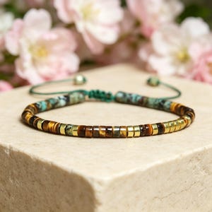 May include: A beaded bracelet with a mix of brown, gold, and turquoise beads. The bracelet is strung on a green cord and is displayed on a light-colored surface with pink flowers in the background.