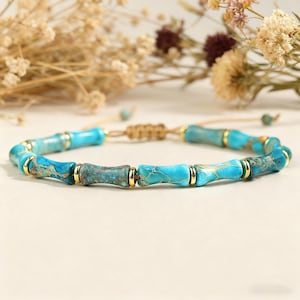 May include: A turquoise beaded bracelet with gold accents. The bracelet features cylindrical beads with a bamboo-like design, connected by a tan cord. The beads are a mix of light and dark turquoise with gold-colored metal rings.