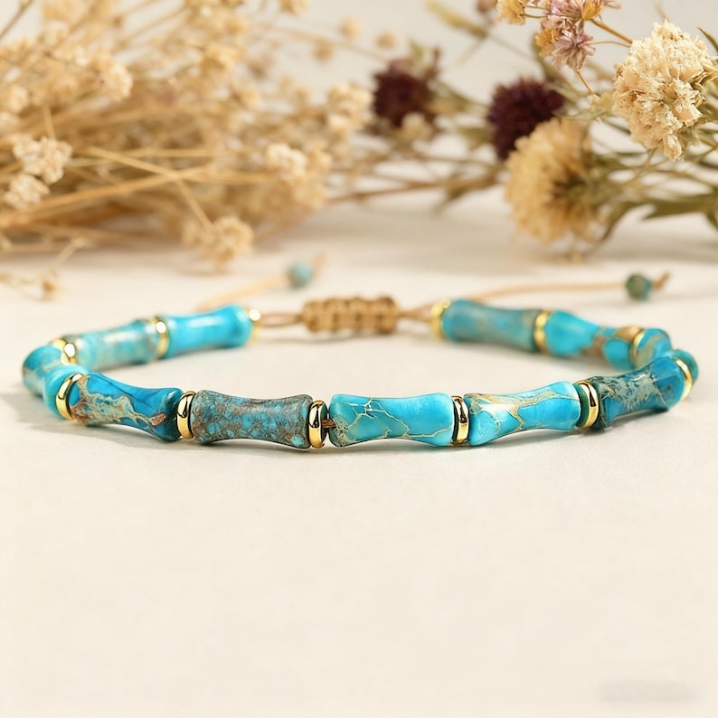 May include: A turquoise beaded bracelet with gold accents. The bracelet features cylindrical beads with a bamboo-like design, connected by a tan cord. The beads are a mix of light and dark turquoise with gold-colored metal rings.