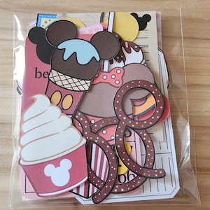 May include: A clear plastic bag filled with colorful paper cutouts. The cutouts feature ice cream cones, cupcakes, and pretzels, all with a Mickey Mouse theme. The word "be" is visible on one of the cutouts. The overall theme is a party or celebration.