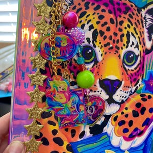 May include: A colorful notebook with a leopard design and a gold star trim. A gold chain with charms, including a pink bead, a green bead, and a charm that says "Sank" in a rainbow font, hangs from the top.