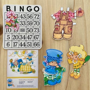 May include: A collection of vintage Strawberry Shortcake themed paper cutouts and a bingo card. The bingo card has the word "BINGO" at the top and numbers in the grid. The cutouts include characters in various outfits and poses.