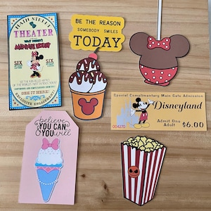 May include: A collection of Disney-themed paper cutouts, including a Minnie Mouse head, ice cream, popcorn, and a Disneyland ticket. The cutouts feature text like "Main Street Theater" and "Believe You Can & You Will."