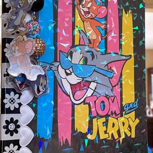 May include: A holographic notebook featuring Tom and Jerry with vertical pink, blue, and yellow stripes. The cover displays Tom wearing blue sunglasses with Jerry standing on his head. A gold-toned charm chain with acrylic character pendants hangs from the side, alongside a black and white spine decorated with flower and heart patterns.