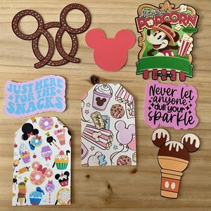 May include: A collection of colorful Disney-themed cutouts and tags. Includes a Mickey Mouse pretzel shape, a pink Mickey head, a Main Street Popcorn Company sign, and tags with text that reads "Just Here For The Snacks" and "Never let anyone dull your sparkle."
