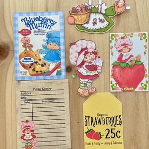 May include: A collection of vintage-style paper cutouts and ephemera. Includes a blueberry muffin mix box, a library card, and several Strawberry Shortcake-themed illustrations. A yellow tag advertises organic strawberries for 25 cents.