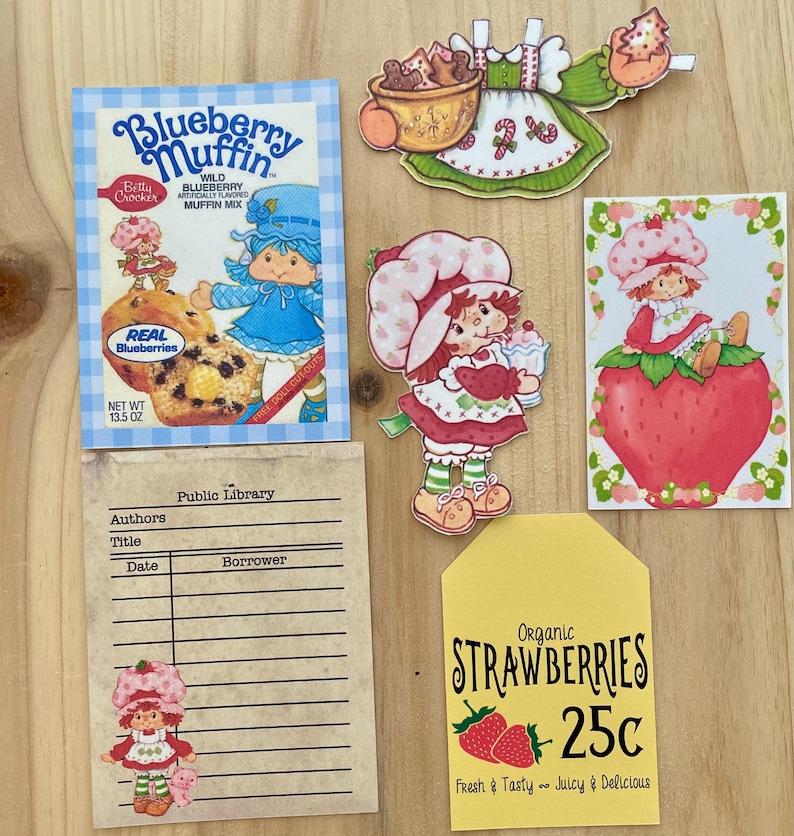 May include: A collection of vintage-style paper cutouts and ephemera. Includes a blueberry muffin mix box, a library card, and several Strawberry Shortcake-themed illustrations. A yellow tag advertises organic strawberries for 25 cents.
