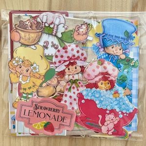 May include: A collection of colorful Strawberry Shortcake themed stickers and cutouts. The images include characters in dresses and bonnets, a pink bathtub, and a sign that reads "Strawberry Lemonade". The stickers are in a clear plastic bag.