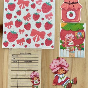 May include: A collection of strawberry-themed paper cutouts and stickers. Includes a pattern of strawberries and bows, a jar of strawberry jam, and several images of a cartoon character in a strawberry hat and dress. A library card is also present.
