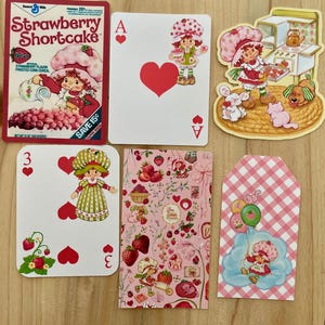 May include: A collection of Strawberry Shortcake themed items. Includes a cereal box, playing cards with heart suits, and decorative tags. The color palette is primarily pink, red, and green, with illustrations of the character and strawberries.