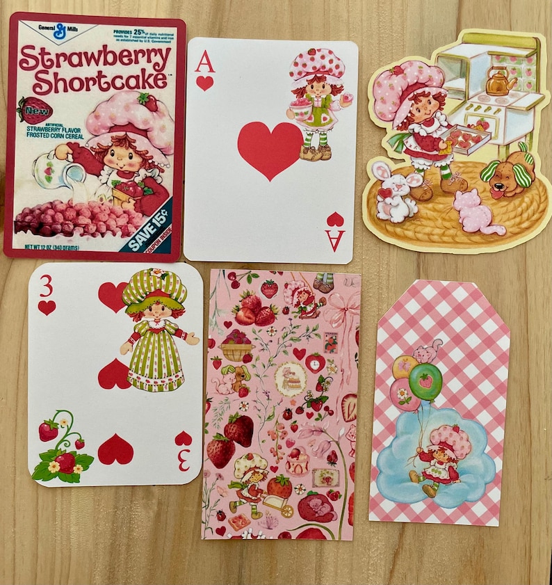 May include: A collection of Strawberry Shortcake themed items. Includes a cereal box, playing cards with heart suits, and decorative tags. The color palette is primarily pink, red, and green, with illustrations of the character and strawberries.