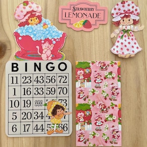 May include: A collection of vintage-style Strawberry Shortcake themed items. Includes a bingo card, a Strawberry Lemonade sign, a patterned paper, and die-cut figures of the character. The color palette is primarily pink, red, and white.