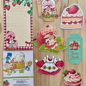 May include: A collection of Strawberry Shortcake themed paper ephemera. Includes a lined notepad, die-cut images of Strawberry Shortcake characters, a cake slice, a cupcake, and a recipe card. The color palette is primarily red, pink, and green.