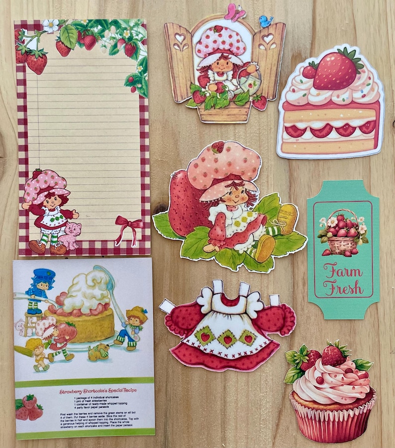 May include: A collection of Strawberry Shortcake themed paper ephemera. Includes a lined notepad, die-cut images of Strawberry Shortcake characters, a cake slice, a cupcake, and a recipe card. The color palette is primarily red, pink, and green.