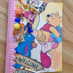 May include: A notebook featuring Winnie the Pooh, Piglet, Eeyore, and Tigger with a 'WELCOME HUNDRED' sign. The spine has a pink heart-patterned trim and a gold-tone metal clip with colorful beads, charms, and character pendants attached.