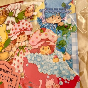 May include: A collection of colorful Strawberry Shortcake stickers. The stickers feature various characters, including one in a pink bathtub with bubbles and a pink cat. Other characters are wearing strawberry-themed hats and dresses.