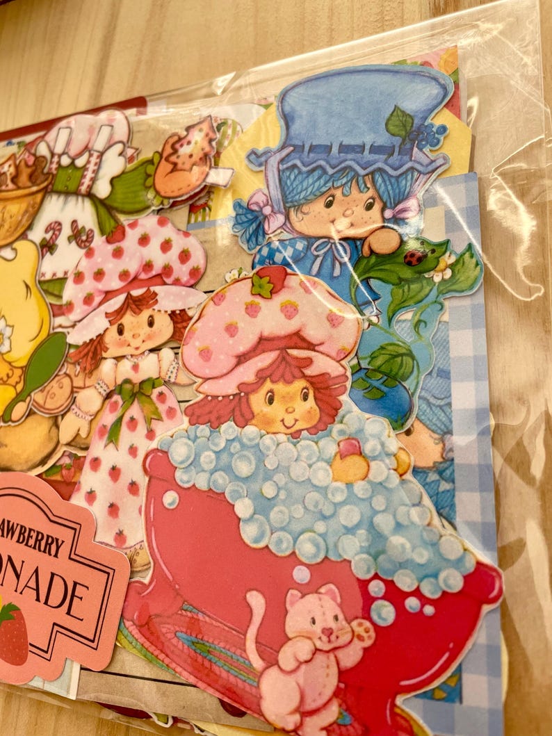 May include: A collection of colorful Strawberry Shortcake stickers. The stickers feature various characters, including one in a pink bathtub with bubbles and a pink cat. Other characters are wearing strawberry-themed hats and dresses.